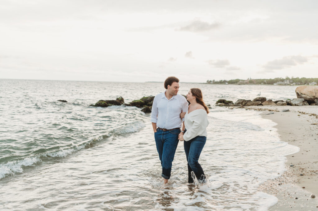 A woman hold onto a man's arm as they look at each other with affection and walk through the water's edge