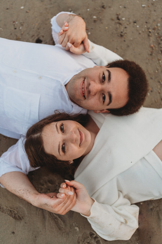 a couple holds hands laying on their backs and looking up at the camera from the sandy beach