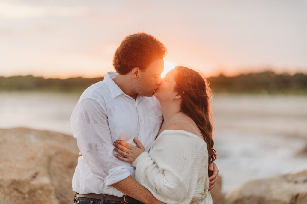 A couple embraces with a romantic kiss and beautiful sunset sun flare in the background at Harkness memorial state park