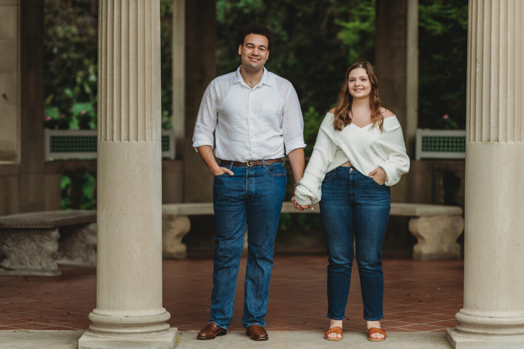 A couple wearing blue jeans and white tops holds hands and looks at the camera from the stone architecture of Harkness Memorial Park