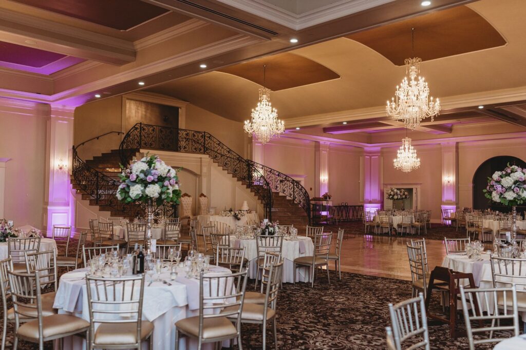A large ballroom with expansive ceiling, large chandeliers, a double staircase, and beautiful flowers and purple uplighting