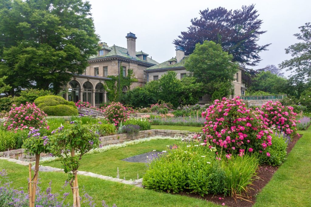 the luscious gardens filled with pink flowers and white and purple blossoms which are well cared for with the eolia mansion at harkness memorial state park in the background.