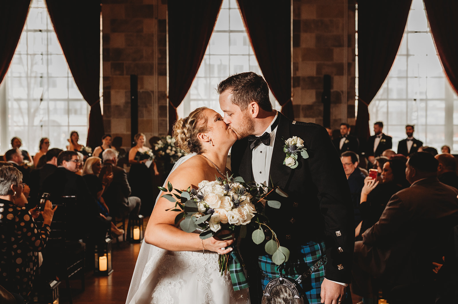 bride and groom in a kilt share a kiss as they walk back up the aisle and the bride holds a white bouquet