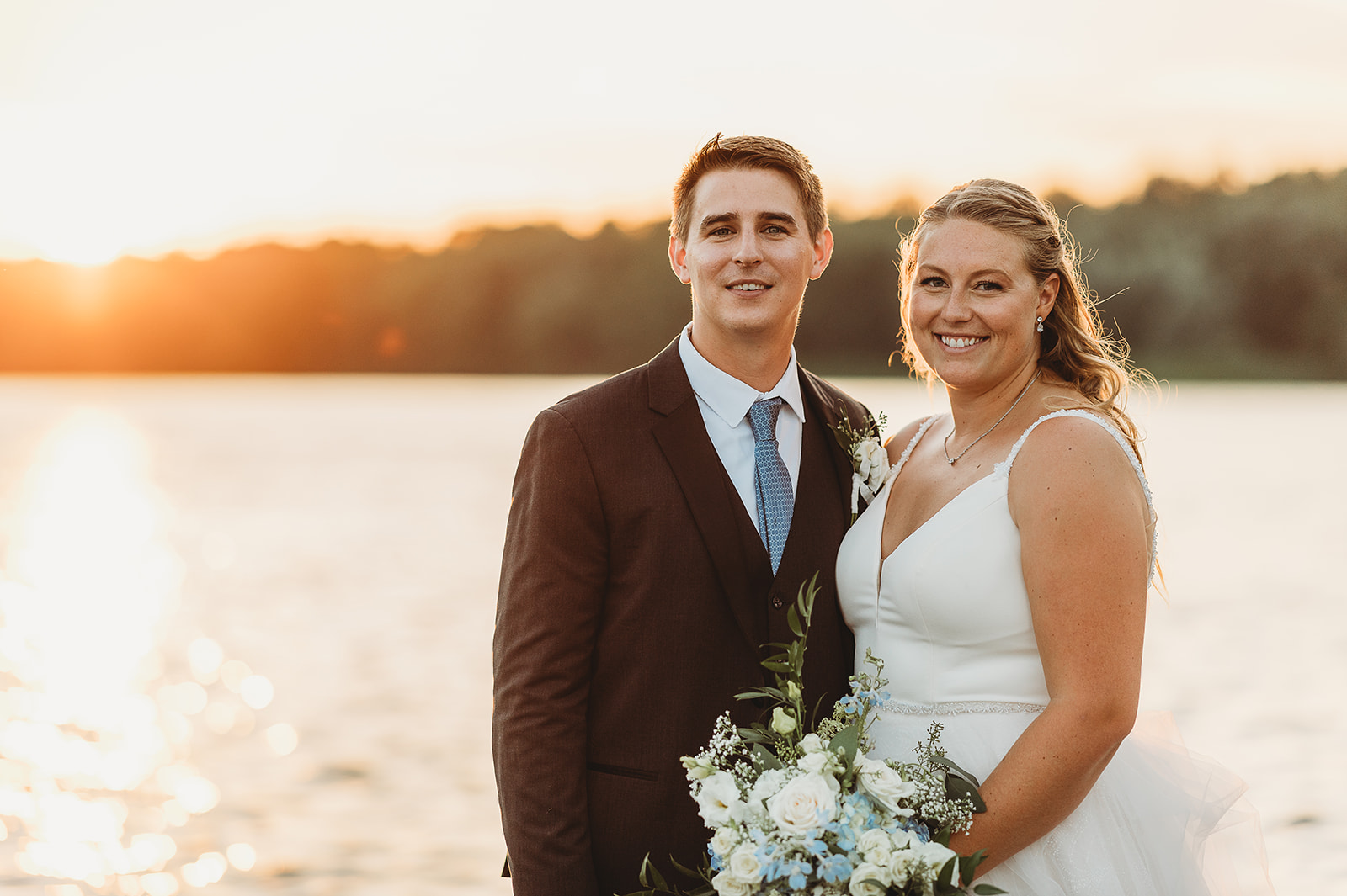 A happy bride and groom smile together while standing by the water at sunset holding a white and blue bouquet