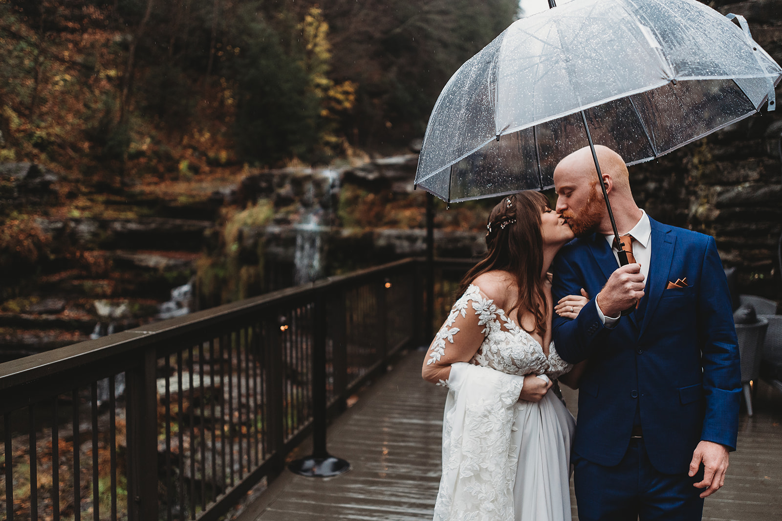 Newlyweds kiss under a clear umbrella on a deck overlooking a waterfall at one of the intimate wedding venues in CT