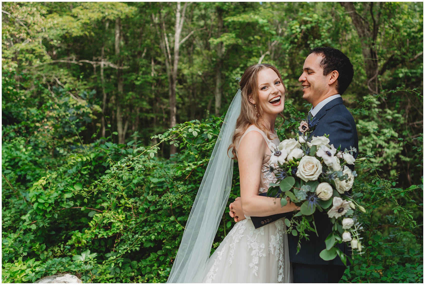 A bride and groom laugh while hugging by the forest holding a large bouquet
