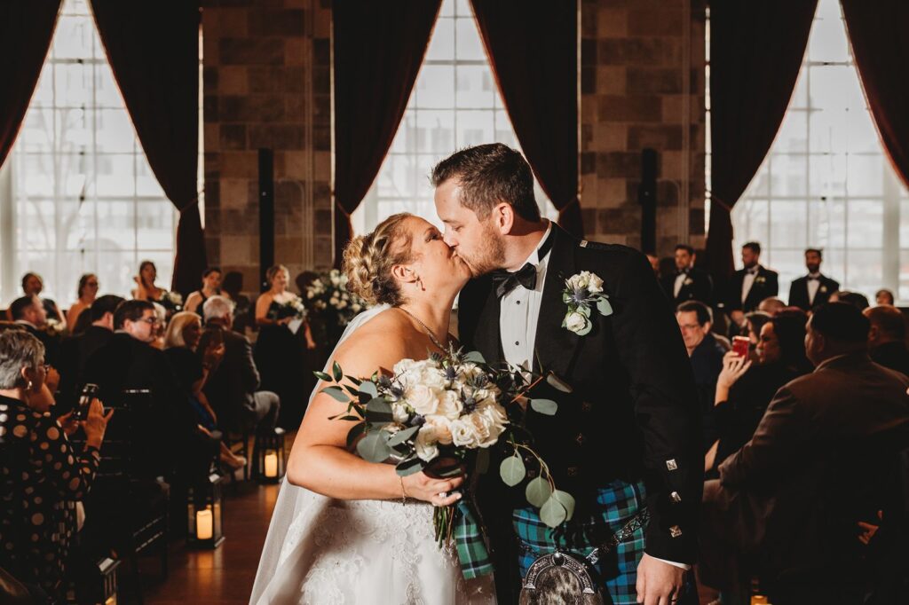 bride holding bouquet and groom wearing kilt kiss on wedding aisle