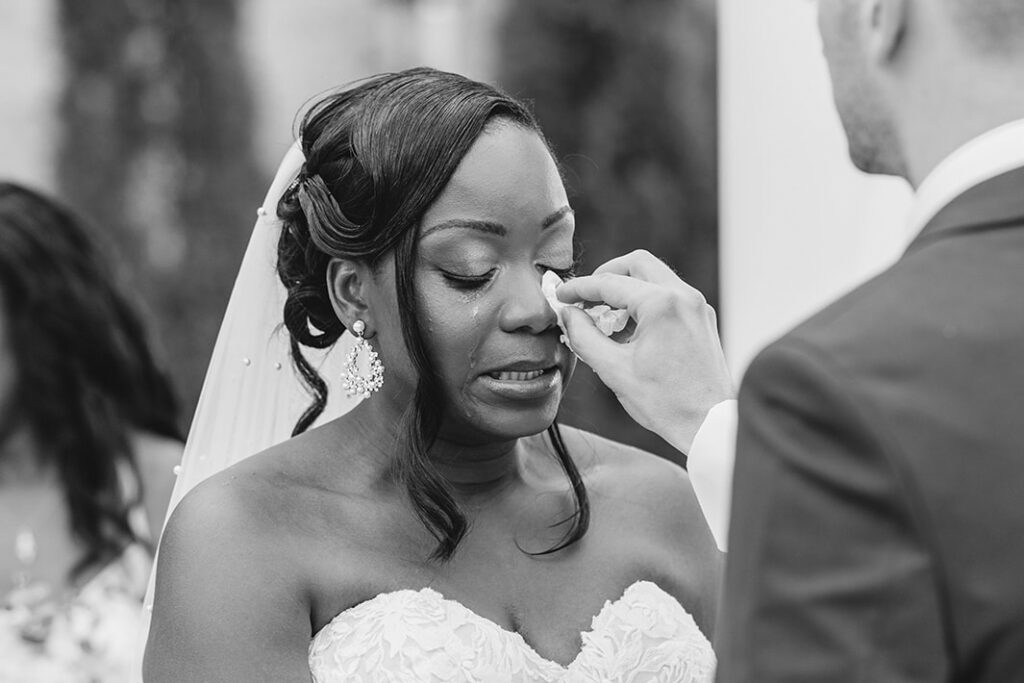 Bride is crying with tears running down her face at the ceremony and the groom wipes her tears with a tissue.
