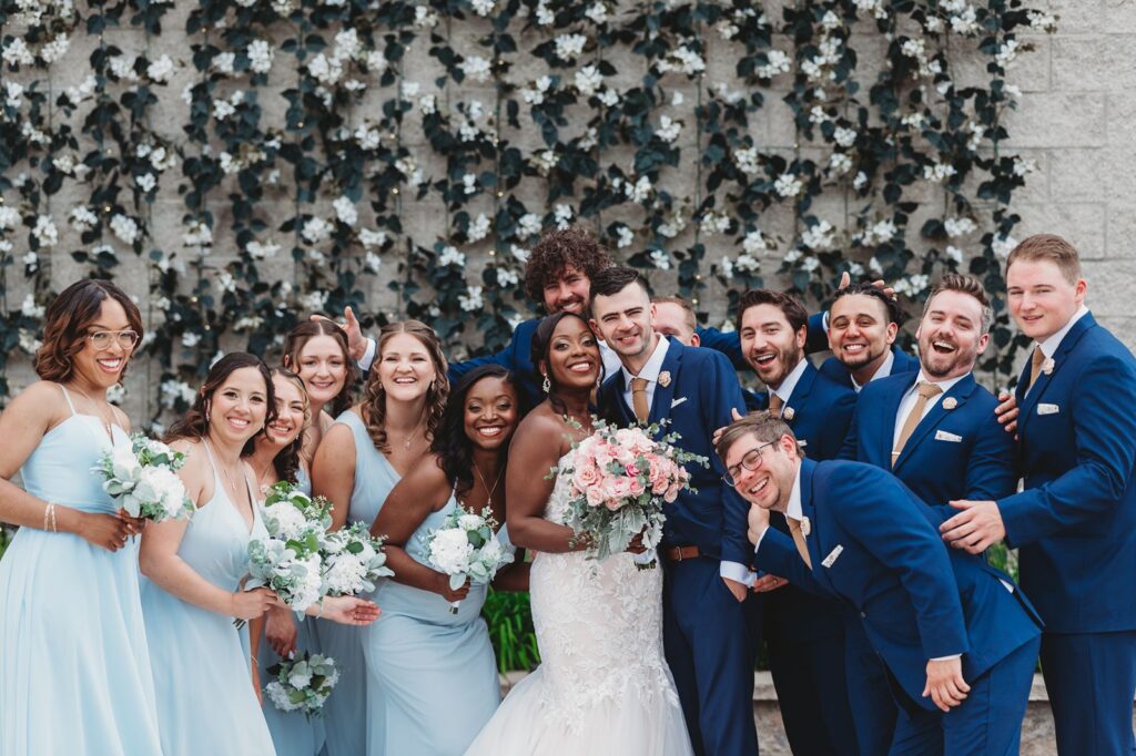 a full wedding party crowds around the bride and groom smiling for a group photo in front of the ivy wall.