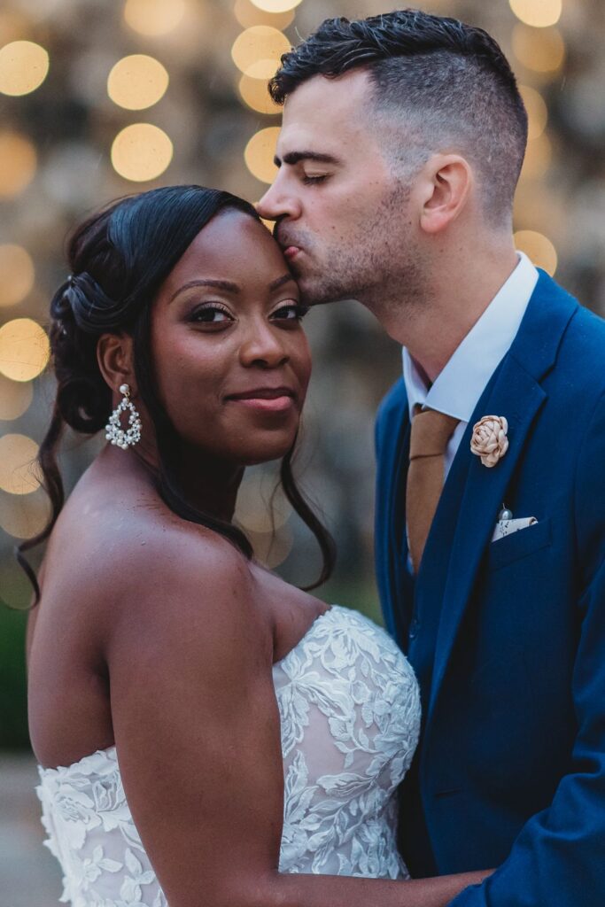 groom kisses bride on side of her head with big lights behind them