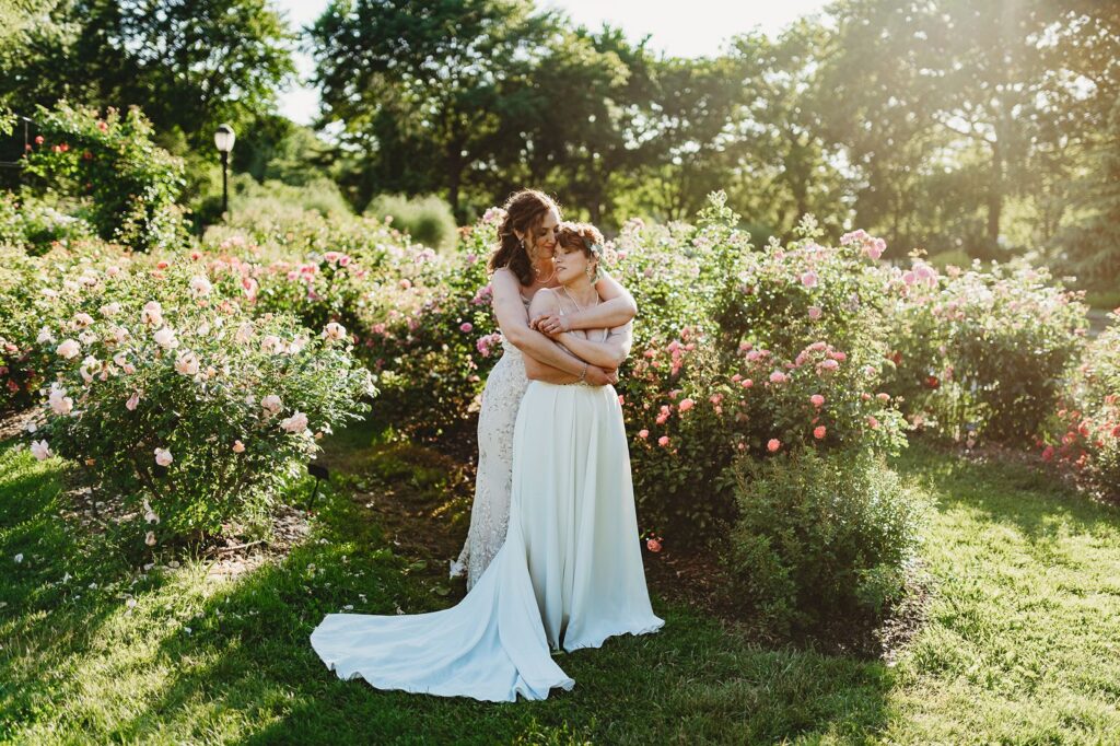 brides embracing in their white wedding dresses in the rose garden at Queens Botanical Gardens